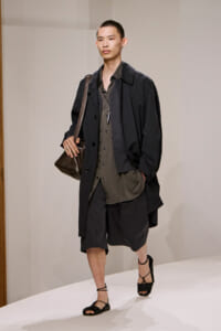 Model walking a runway in a loose black coat over an olive button-down, black knee-length shorts, strappy black sandals, and a brown crossbody bag.