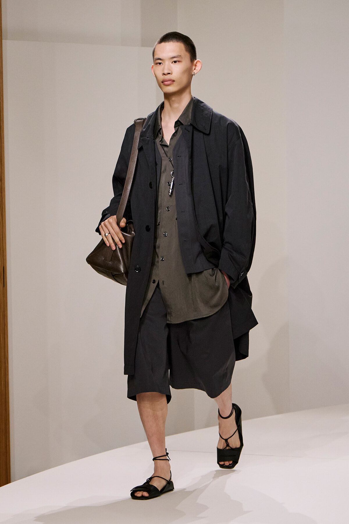 Model walking a runway in a loose black coat over an olive button-down, black knee-length shorts, strappy black sandals, and a brown crossbody bag.