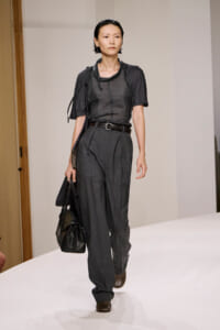 Model walks down the runway in a dark sheer top tucked into gray high-waisted trousers with a brown belt, carrying a black bag.