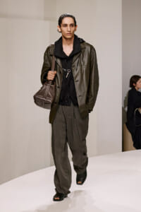 Male model walking a runway in an olive leather jacket, dark layers, and dark bag slung over his shoulder with open-toe sandals.