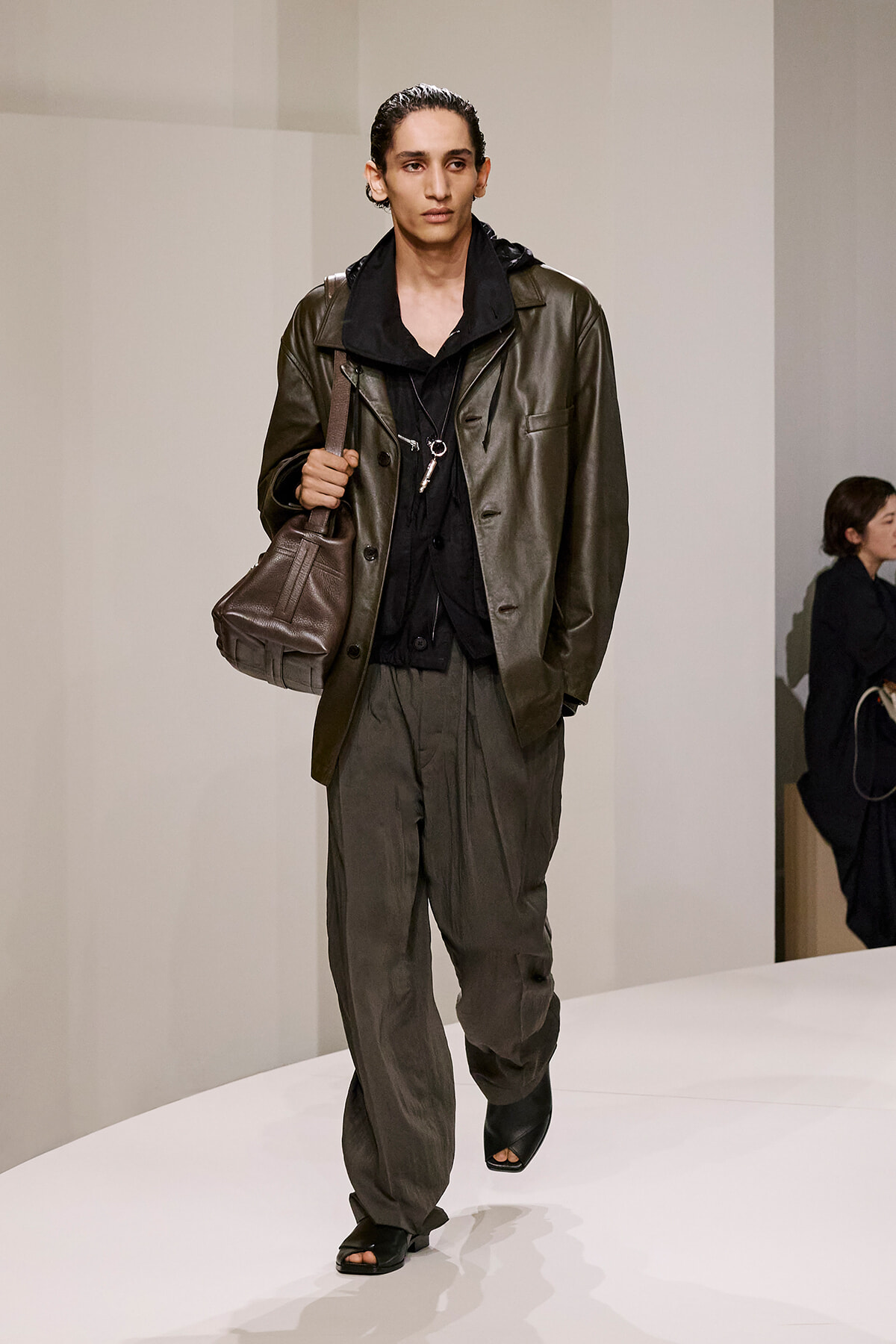 Male model walking a runway in an olive leather jacket, dark layers, and dark bag slung over his shoulder with open-toe sandals.