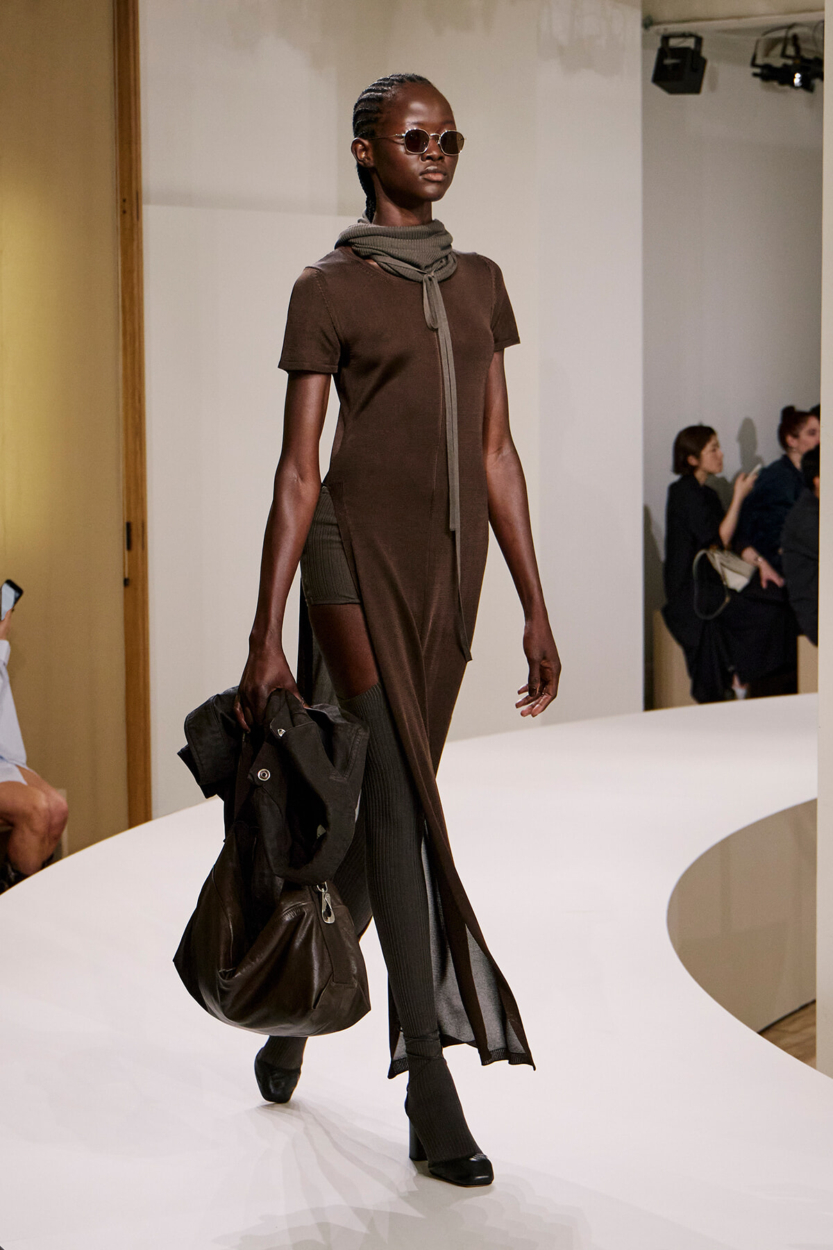 Model walking a curved white runway in a brown midi dress with a scarf, sunglasses, and carrying a dark coat and large bag in one hand.