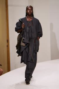 Model walking a fashion show in a loose, dark gray utility jumpsuit with oversized sleeves and multiple pockets, carrying a black bag over the shoulder.