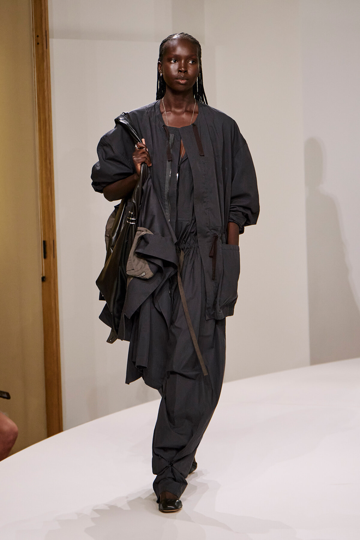 Model walking a fashion show in a loose, dark gray utility jumpsuit with oversized sleeves and multiple pockets, carrying a black bag over the shoulder.