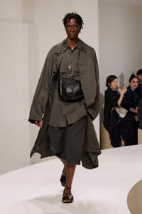 Male runway model wearing an oversized olive-gray trench coat over a matching shirt and baggy shorts, with a black leather waist pouch and layered necklaces as he strides down a white catwalk.