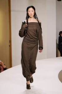 Model walks a runway in a brown off-shoulder knit jumpsuit with cuffed ankles and brown mules.