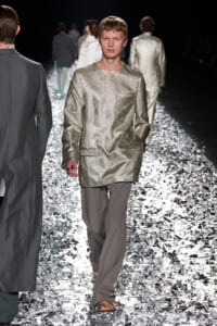 Male model in a metallic gold long-sleeve tunic and gray trousers walks the glittering runway during a fashion show.