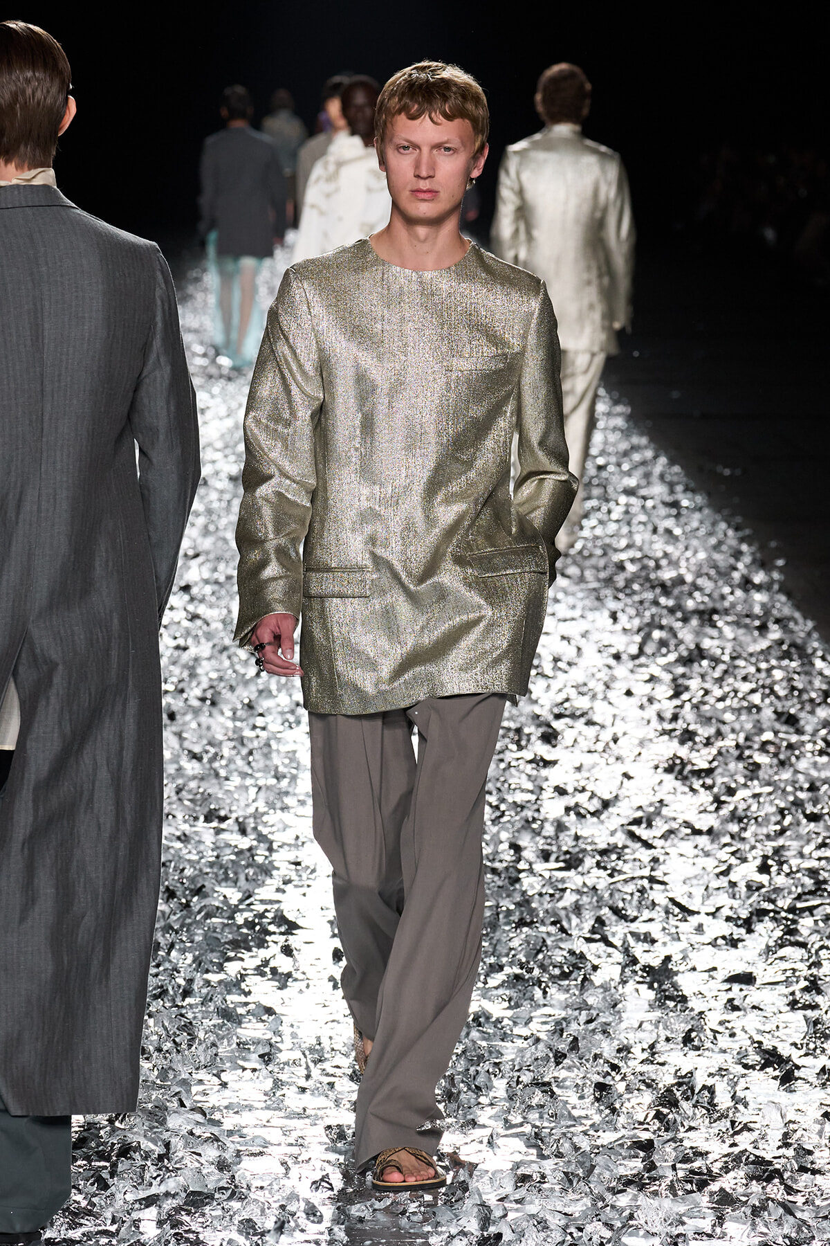 Male model in a metallic gold long-sleeve tunic and gray trousers walks the glittering runway during a fashion show.