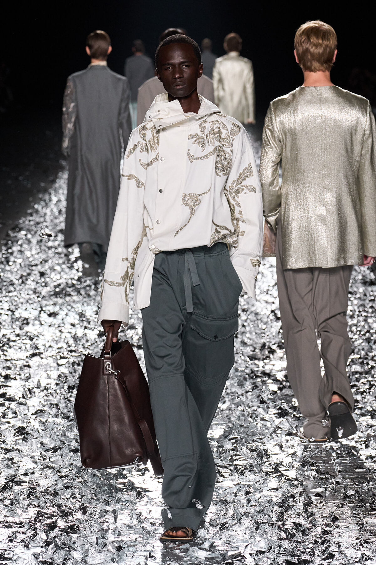 Model walks a fashion show in a white embroidered jacket, dark green cargo pants, and a brown tote bag, with metallic floor reflection behind.