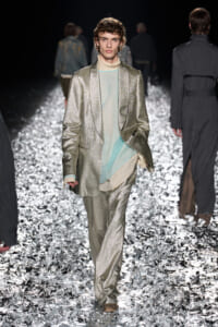 Male model walks the runway in a metallic silver suit with a layered sheer pastel top peeking from underneath, glittering floor beneath.