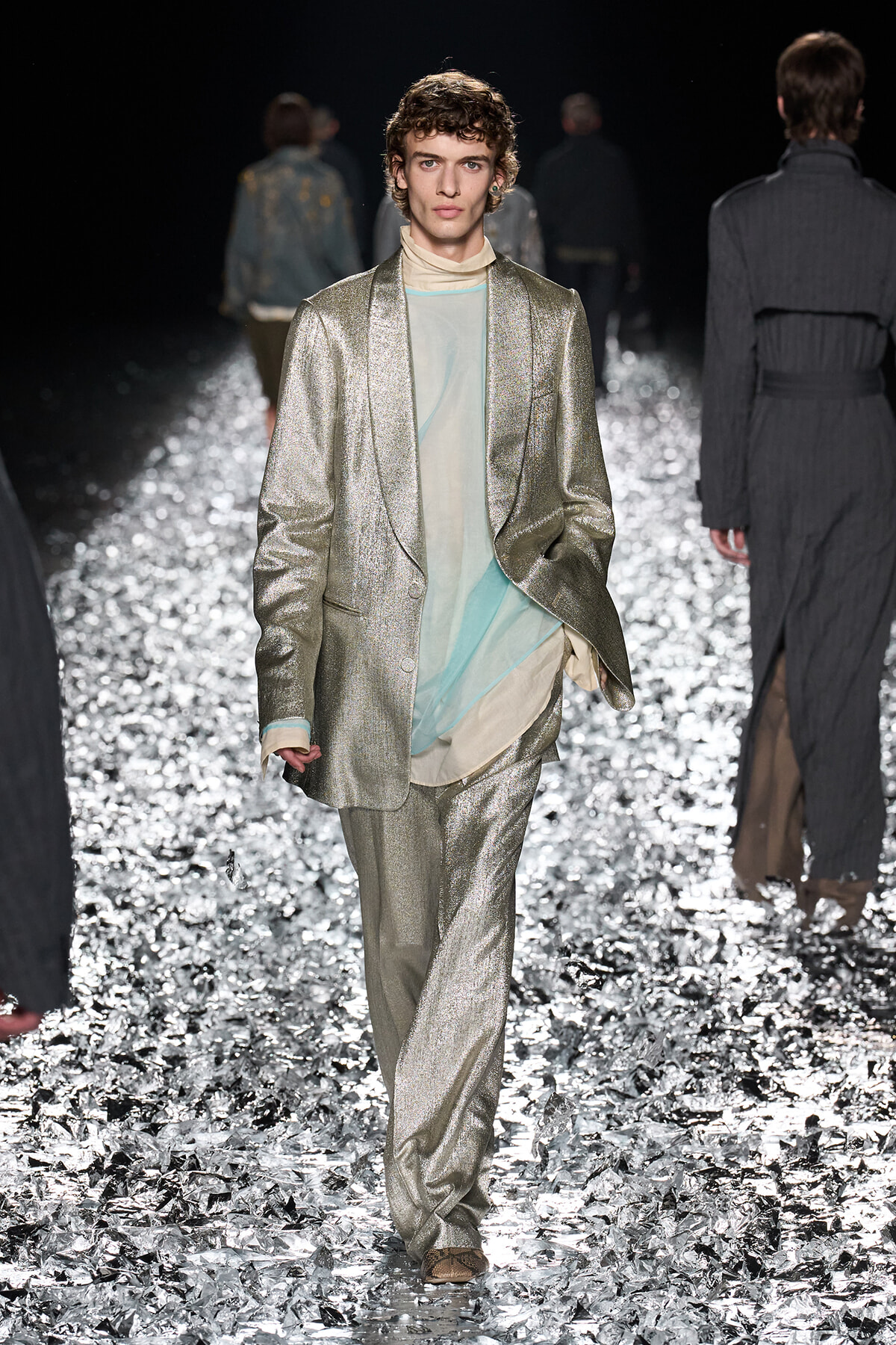 Male model walks the runway in a metallic silver suit with a layered sheer pastel top peeking from underneath, glittering floor beneath.