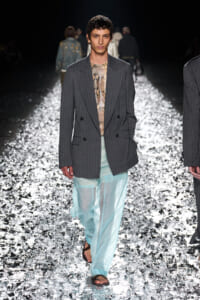 Male model walks a glittery runway in a gray pinstripe blazer, snakeskin top, and sheer light-blue pants, wearing sandals. Runway model wearing an oversized gray pinstripe blazer over a snakeskin top and translucent aqua trousers on a reflective catwalk. Fashion show scene: model in a gray double-breasted blazer and sheer turquoise trousers on a metallic, reflective runway. Model on the catwalk showcasing a dark pinstripe blazer, snakeskin shirt, and airy sheer pants amid a sparkly runway backdrop.