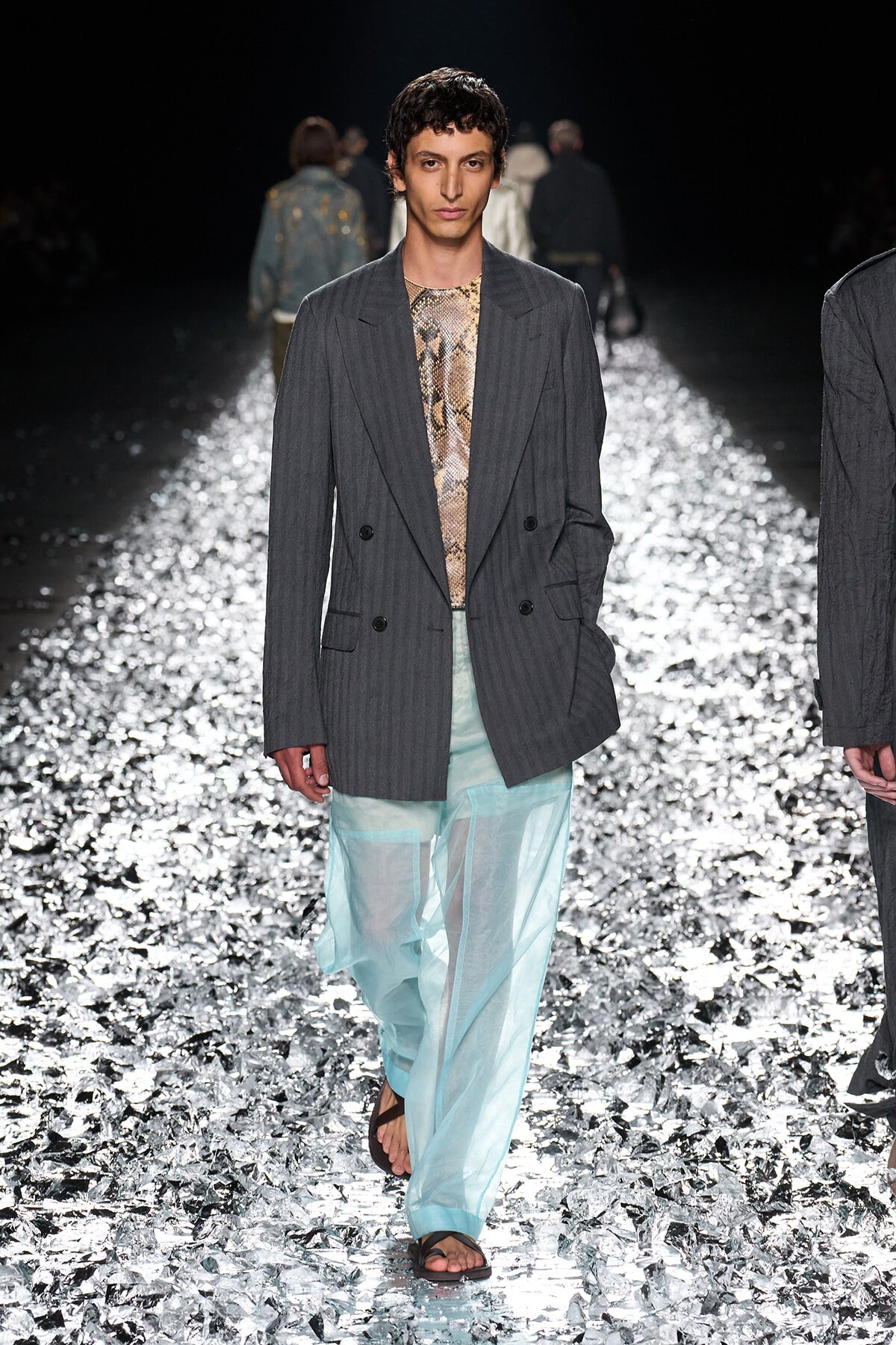 Male model walks a glittery runway in a gray pinstripe blazer, snakeskin top, and sheer light-blue pants, wearing sandals. Runway model wearing an oversized gray pinstripe blazer over a snakeskin top and translucent aqua trousers on a reflective catwalk. Fashion show scene: model in a gray double-breasted blazer and sheer turquoise trousers on a metallic, reflective runway. Model on the catwalk showcasing a dark pinstripe blazer, snakeskin shirt, and airy sheer pants amid a sparkly runway backdrop.