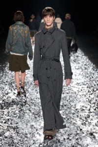 Model walks the runway in a gray pinstriped trench coat with a belt on a glittering, reflective catwalk.