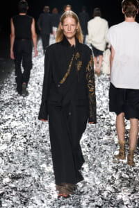 Model walks a metallic foil-covered runway wearing a long black coat with gold floral embroidery on the sleeve and chest.