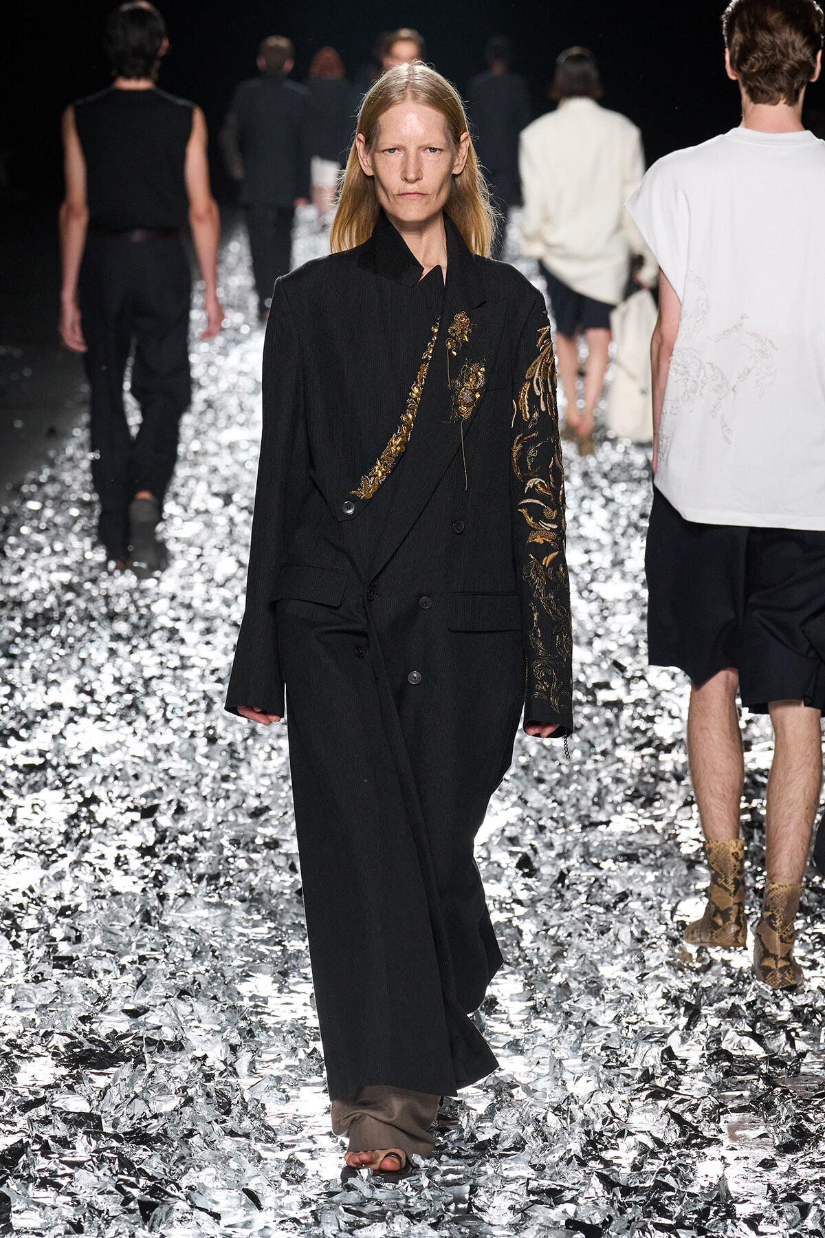 Model walks a metallic foil-covered runway wearing a long black coat with gold floral embroidery on the sleeve and chest.