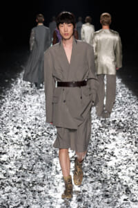 Model walks the runway in a gray oversized blazer-dress cinched with a wide dark belt, paired with snakeskin ankle boots on a glittering metallic catwalk.