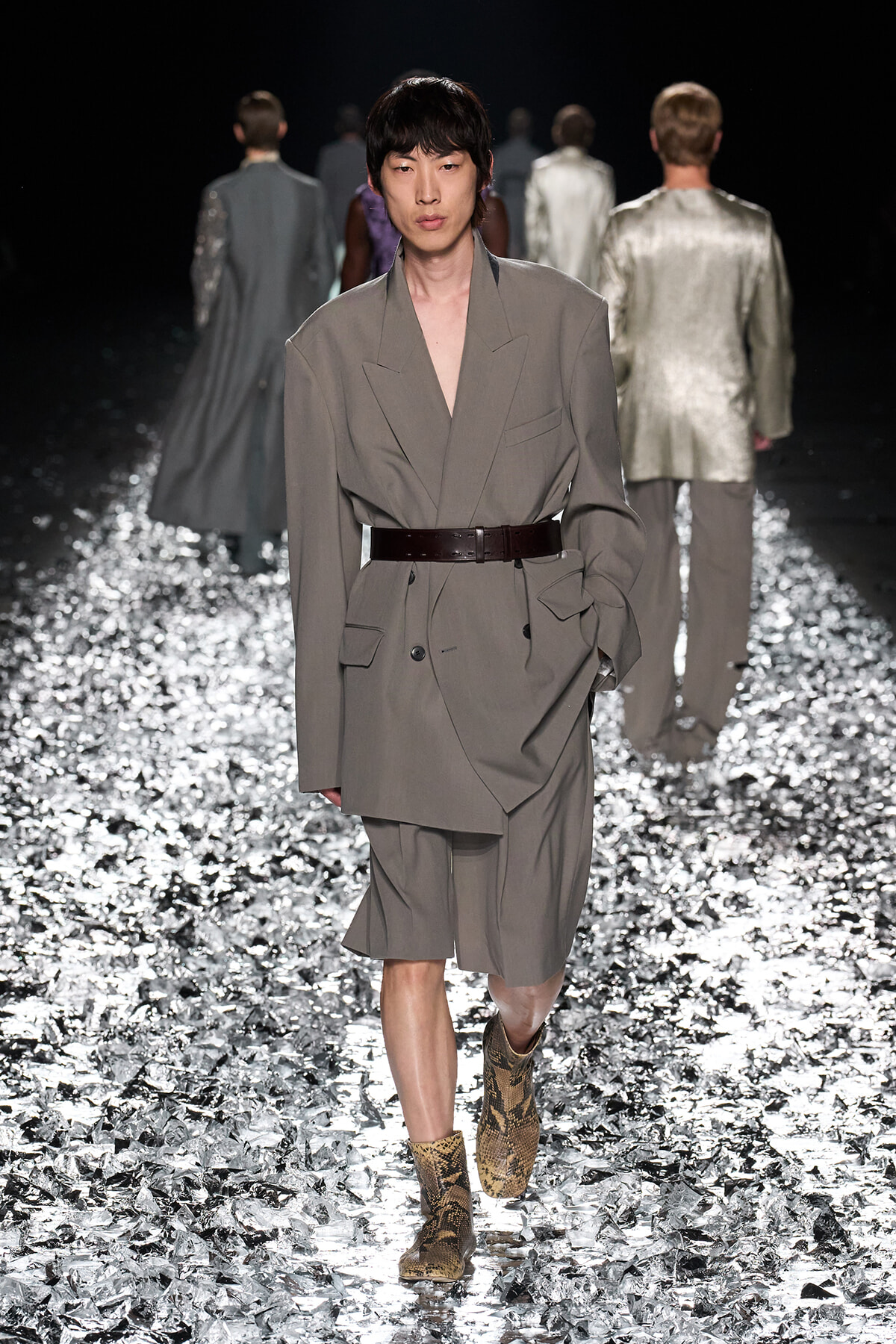 Model walks the runway in a gray oversized blazer-dress cinched with a wide dark belt, paired with snakeskin ankle boots on a glittering metallic catwalk.