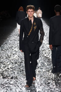 Male model walks a black double-breasted suit with gold embroidery down a reflective, foil-covered runway at a fashion show.