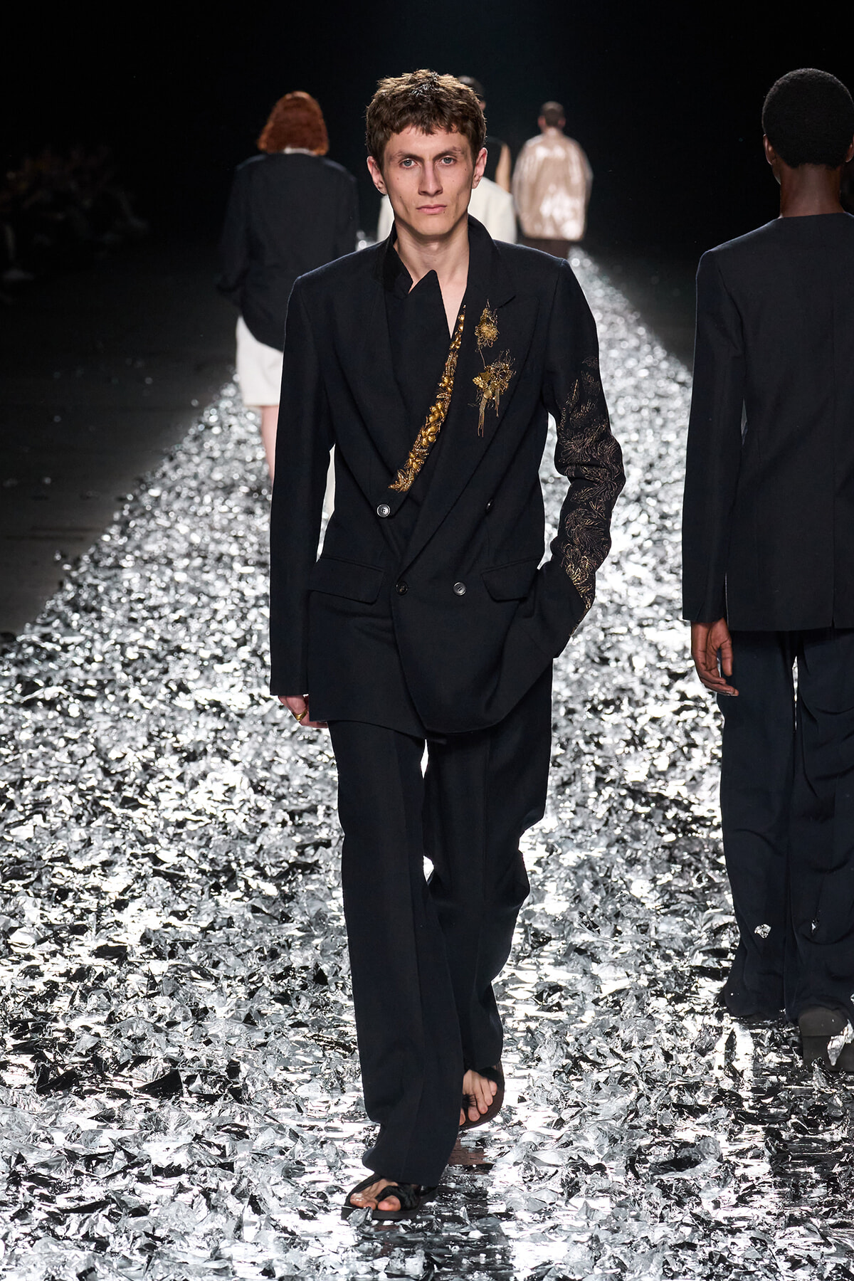 Male model walks a black double-breasted suit with gold embroidery down a reflective, foil-covered runway at a fashion show.