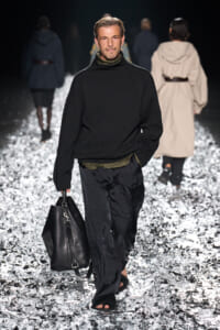 Male model on a fashion runway wearing a black knit turtleneck and olive layer, carrying a black leather duffel bag.