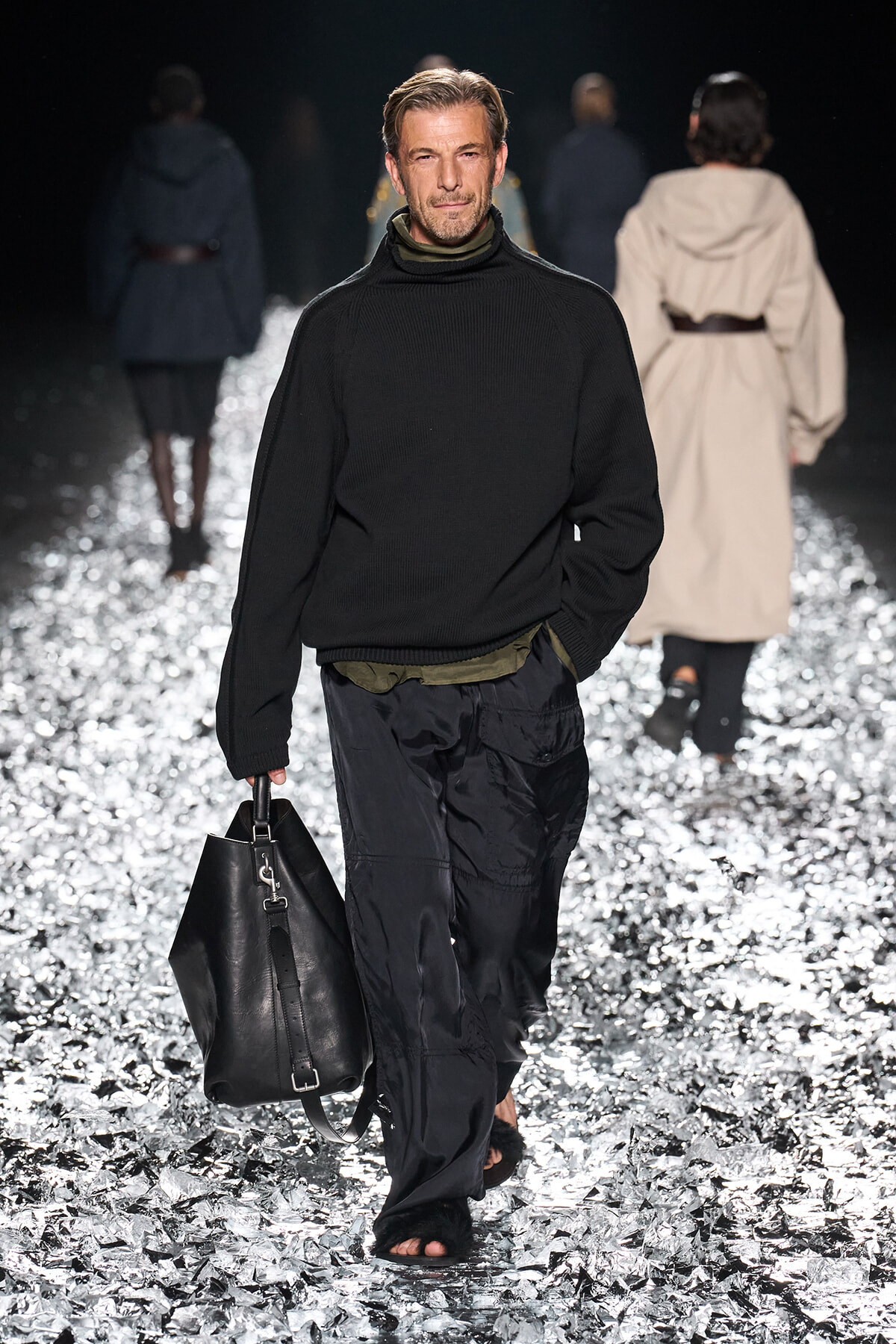 Male model on a fashion runway wearing a black knit turtleneck and olive layer, carrying a black leather duffel bag.