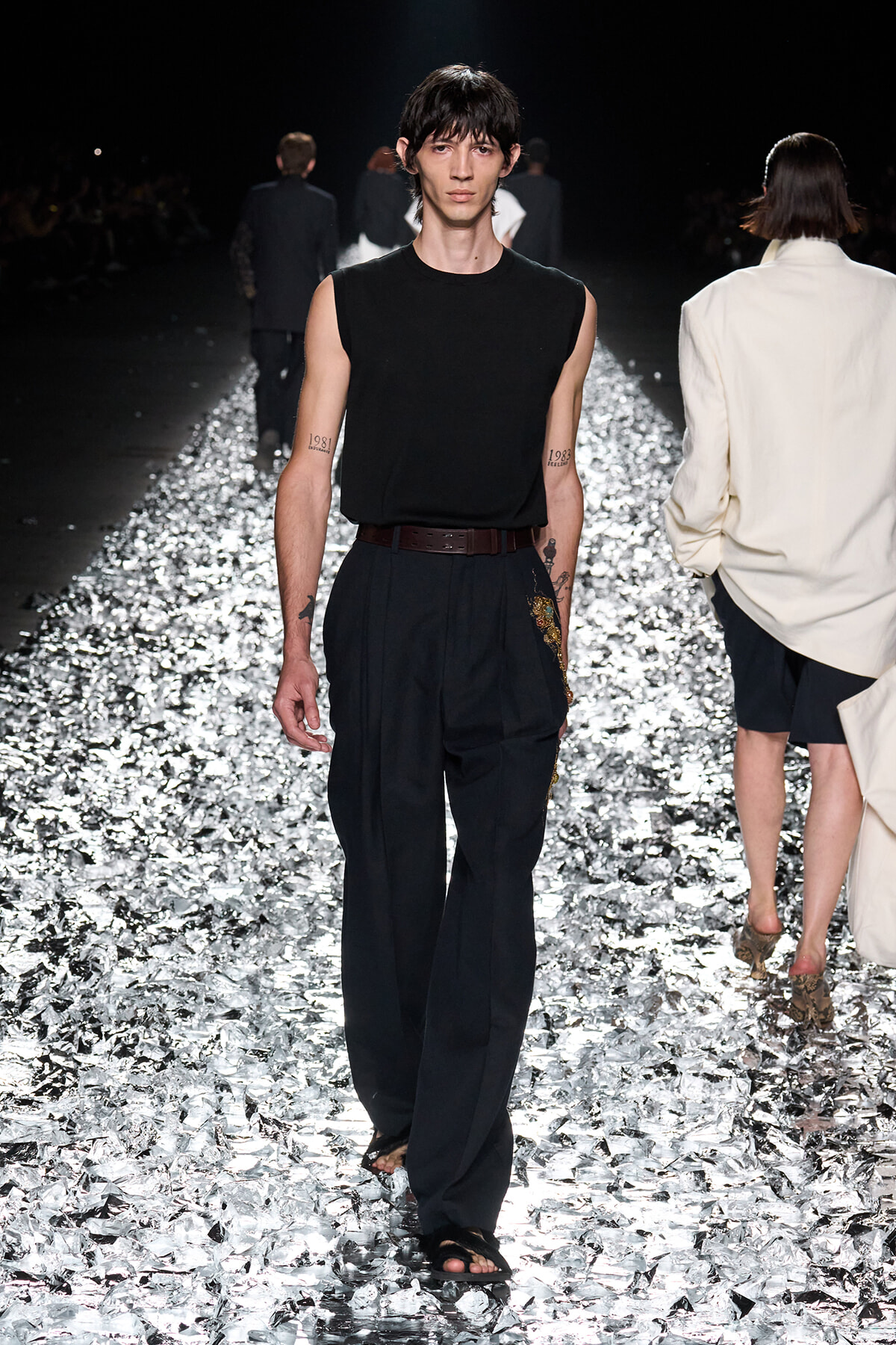 Male model walking a metallic, confetti-covered runway in a black sleeveless top and dark trousers with a brown belt and sandals.