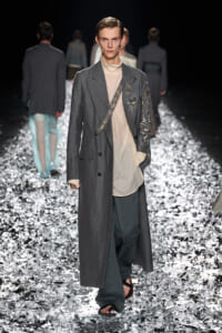 Male model on a glittery runway wears a long gray coat over a beige tunic, with an embroidered strap crossing the chest and ornate sleeve details.