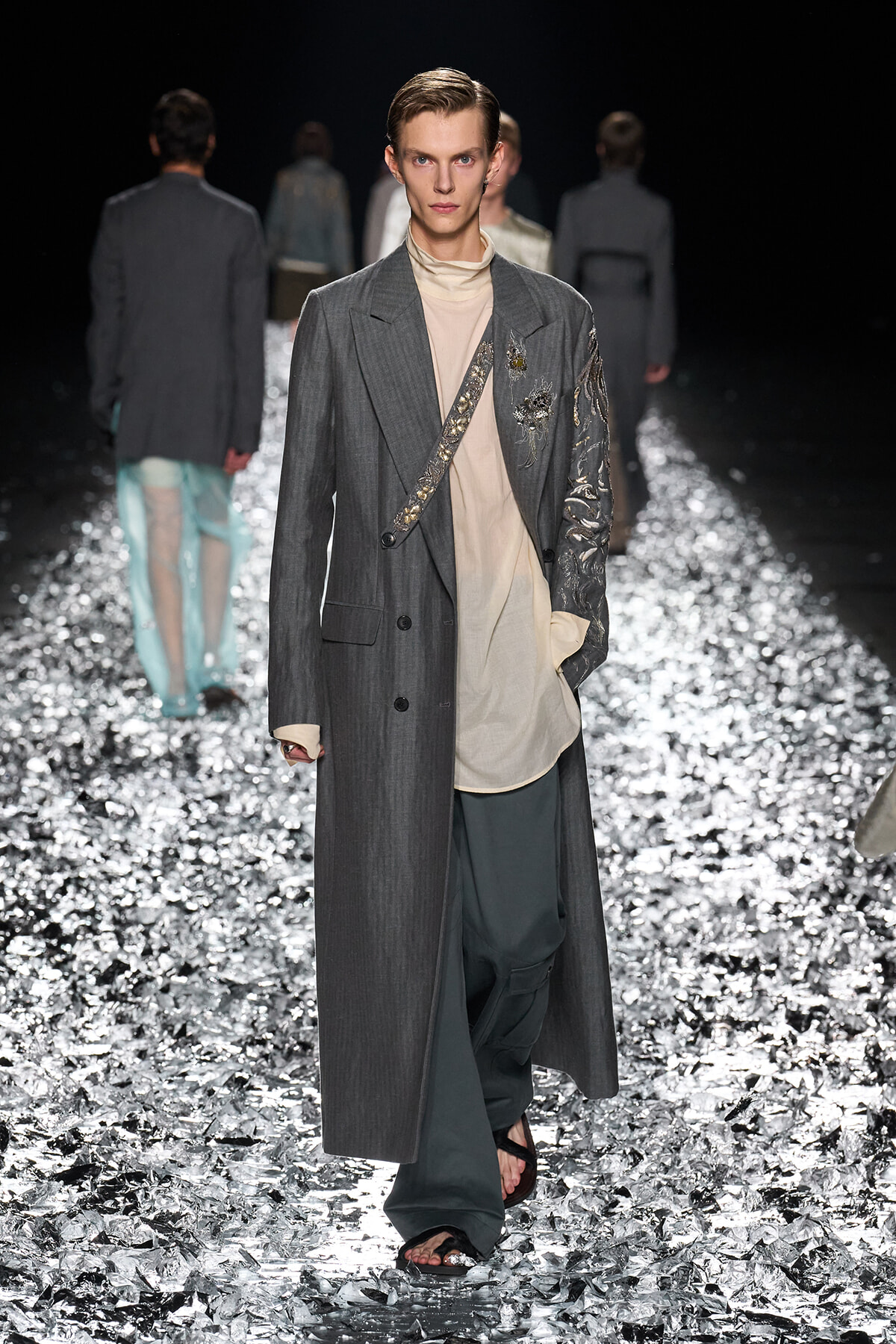 Male model on a glittery runway wears a long gray coat over a beige tunic, with an embroidered strap crossing the chest and ornate sleeve details.