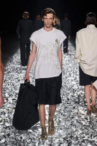 Male fashion model walking a runway in a white printed T-shirt, black knee-length shorts, and snakeskin boots, carrying a large black bag on his side amid a metallic, reflective runway.