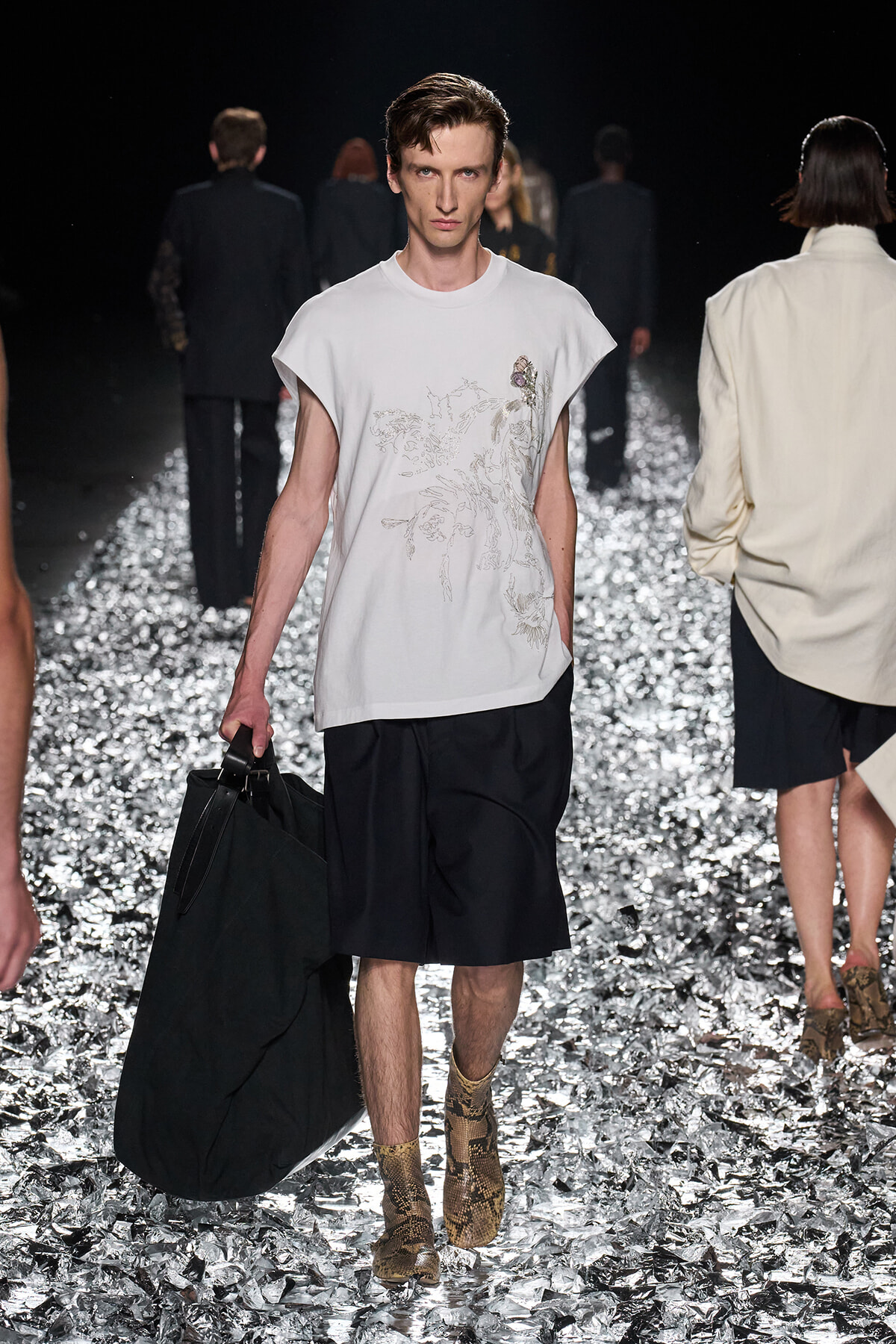 Male fashion model walking a runway in a white printed T-shirt, black knee-length shorts, and snakeskin boots, carrying a large black bag on his side amid a metallic, reflective runway.