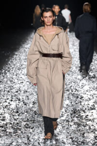 Model walks a fashion runway wearing a beige oversized trench with a wide brown belt, beige top, and black pants, on a glittering metallic runway surface.