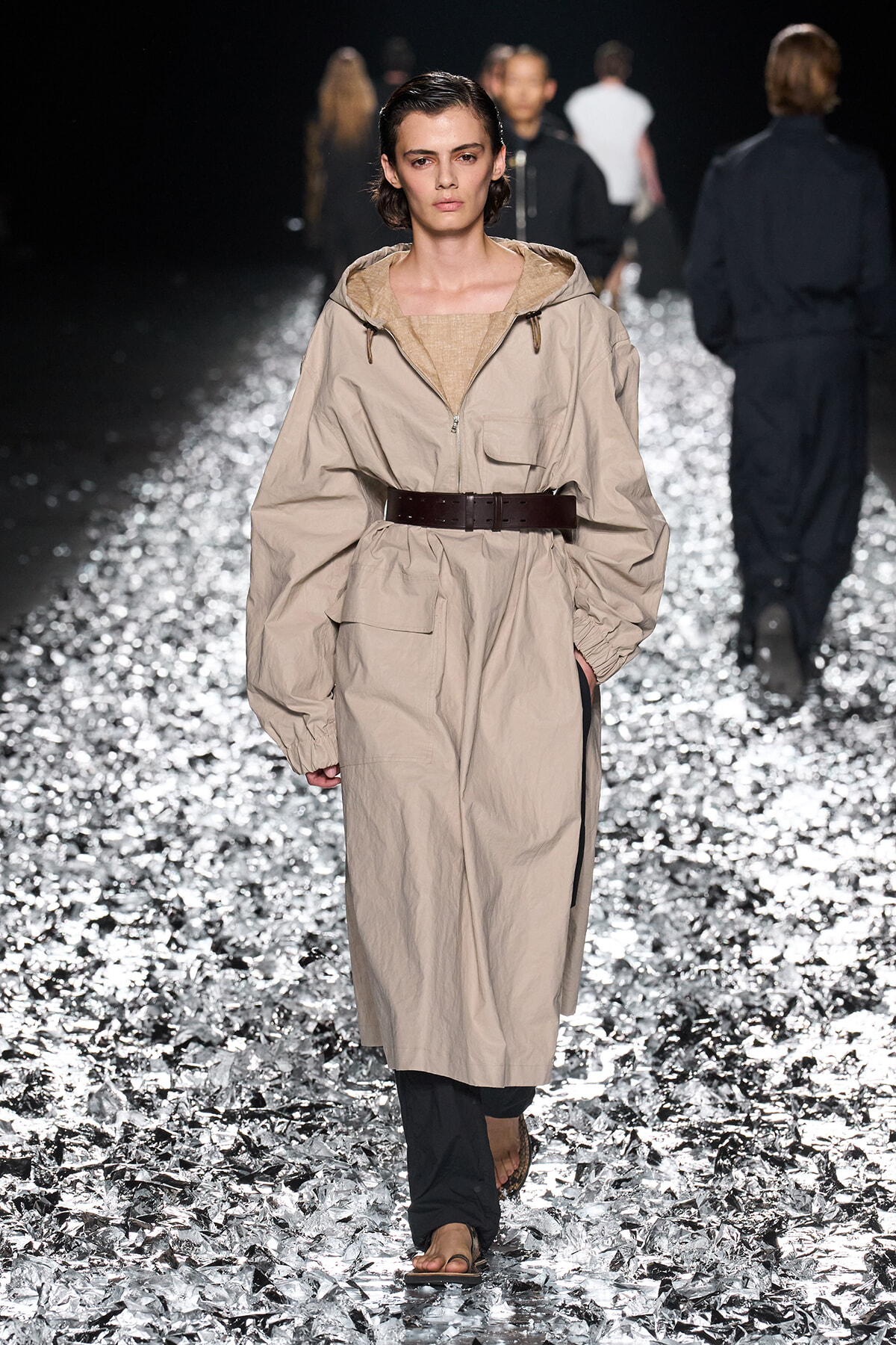 Model walks a fashion runway wearing a beige oversized trench with a wide brown belt, beige top, and black pants, on a glittering metallic runway surface.