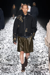 Male model on a glittering runway wearing a black embroidered bomber jacket with gold floral appliqués and olive leather shorts, brown boots visible.