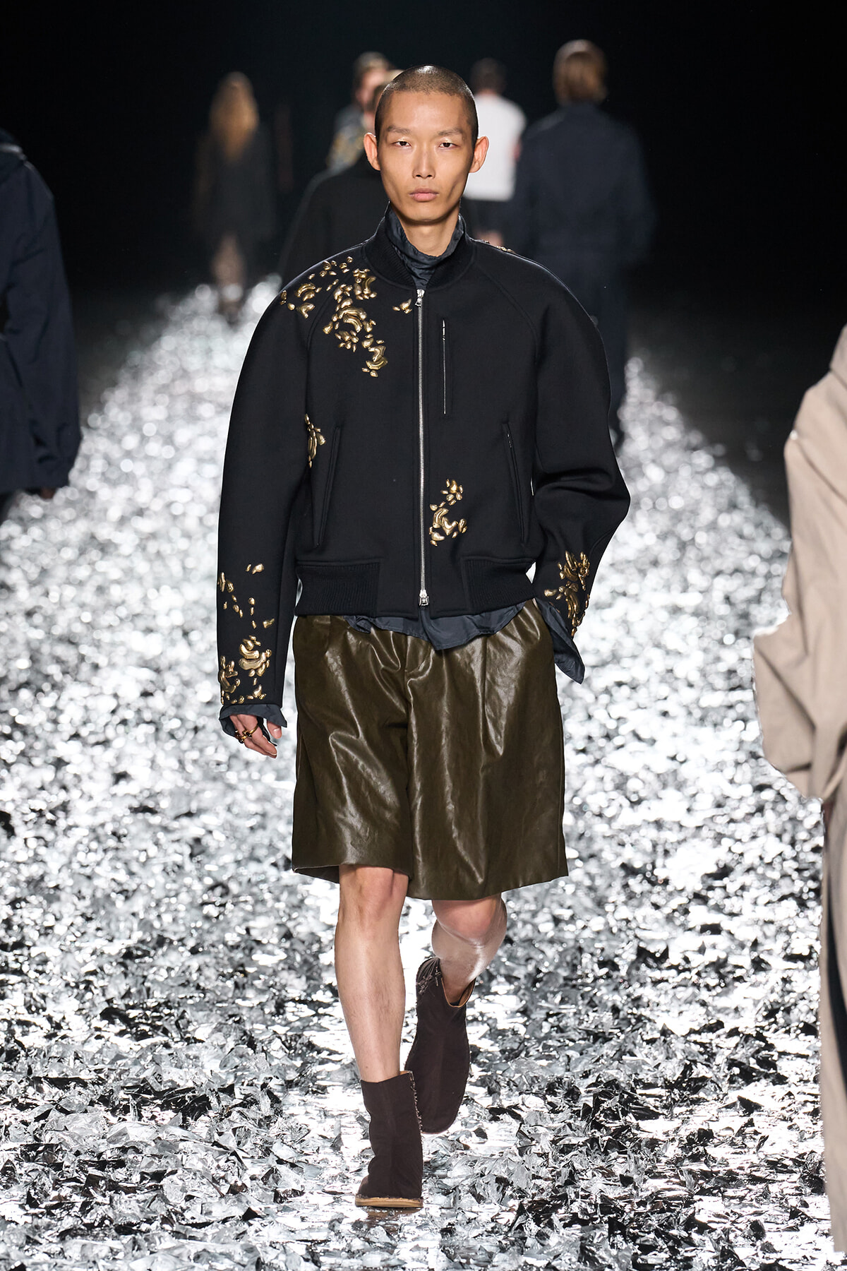 Male model on a glittering runway wearing a black embroidered bomber jacket with gold floral appliqués and olive leather shorts, brown boots visible.