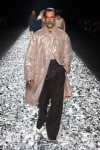 Male model walks the runway in a beige shiny coat over dark pants on a glittering, metallic catwalk.