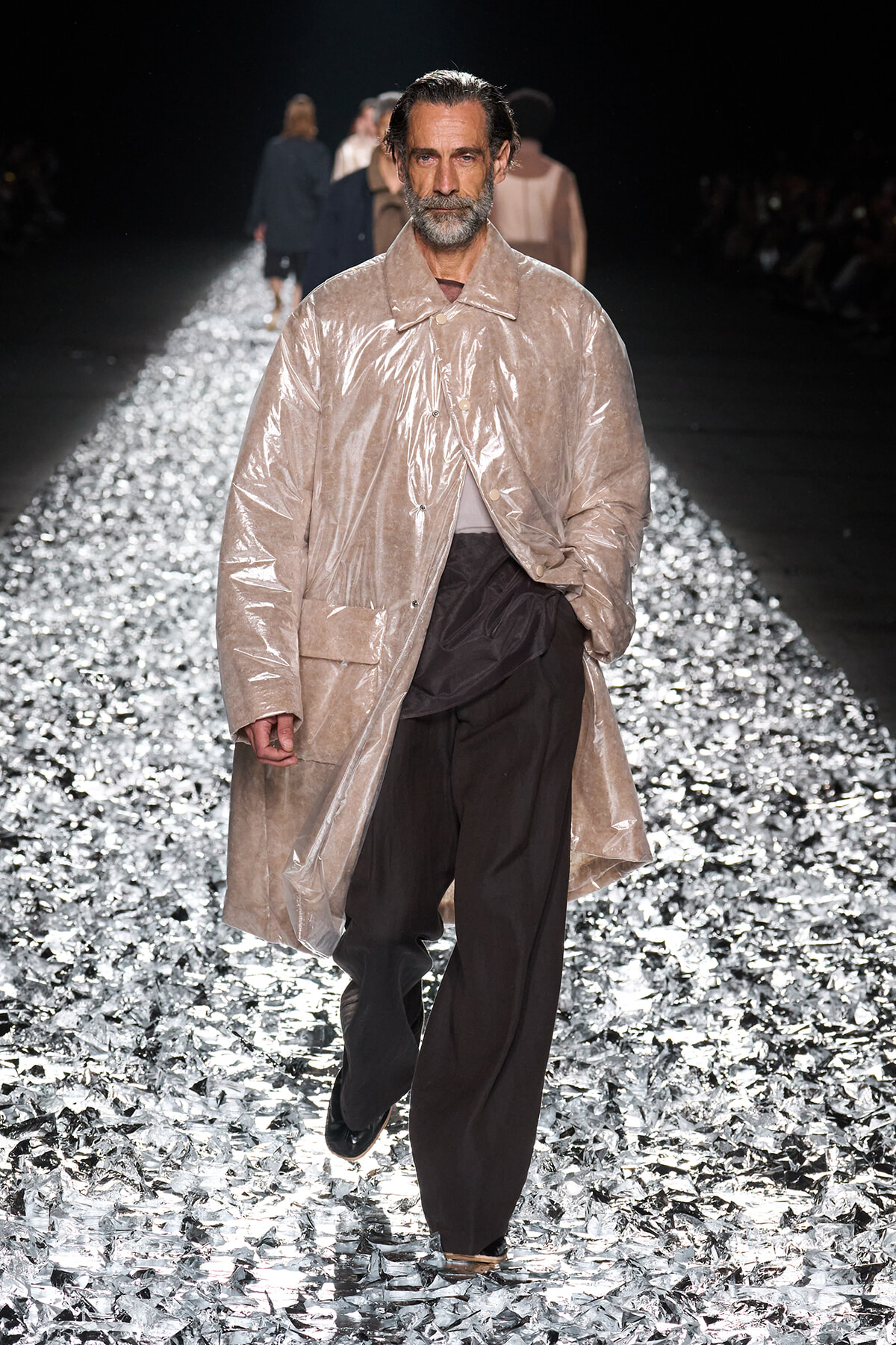 Male model walks the runway in a beige shiny coat over dark pants on a glittering, metallic catwalk.