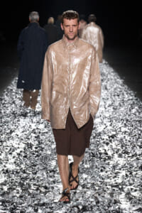 Male model on a fashion runway wearing a shiny beige oversized shirt, brown knee-length shorts, and sandals on a foil-covered catwalk.