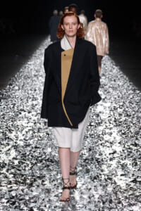 Model with red hair walks a metallic runway wearing a black oversized blazer with a tan inner panel over a light outfit and strappy sandals.