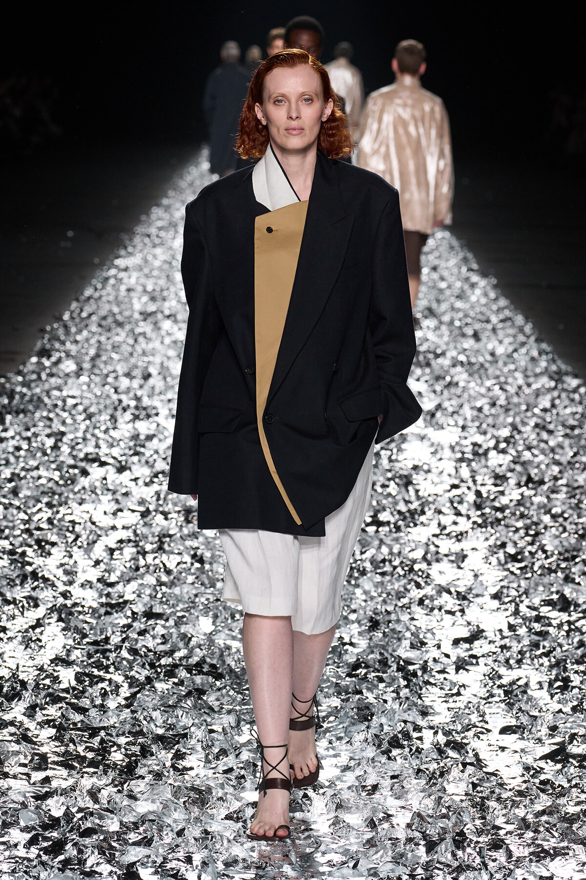 Model with red hair walks a metallic runway wearing a black oversized blazer with a tan inner panel over a light outfit and strappy sandals.