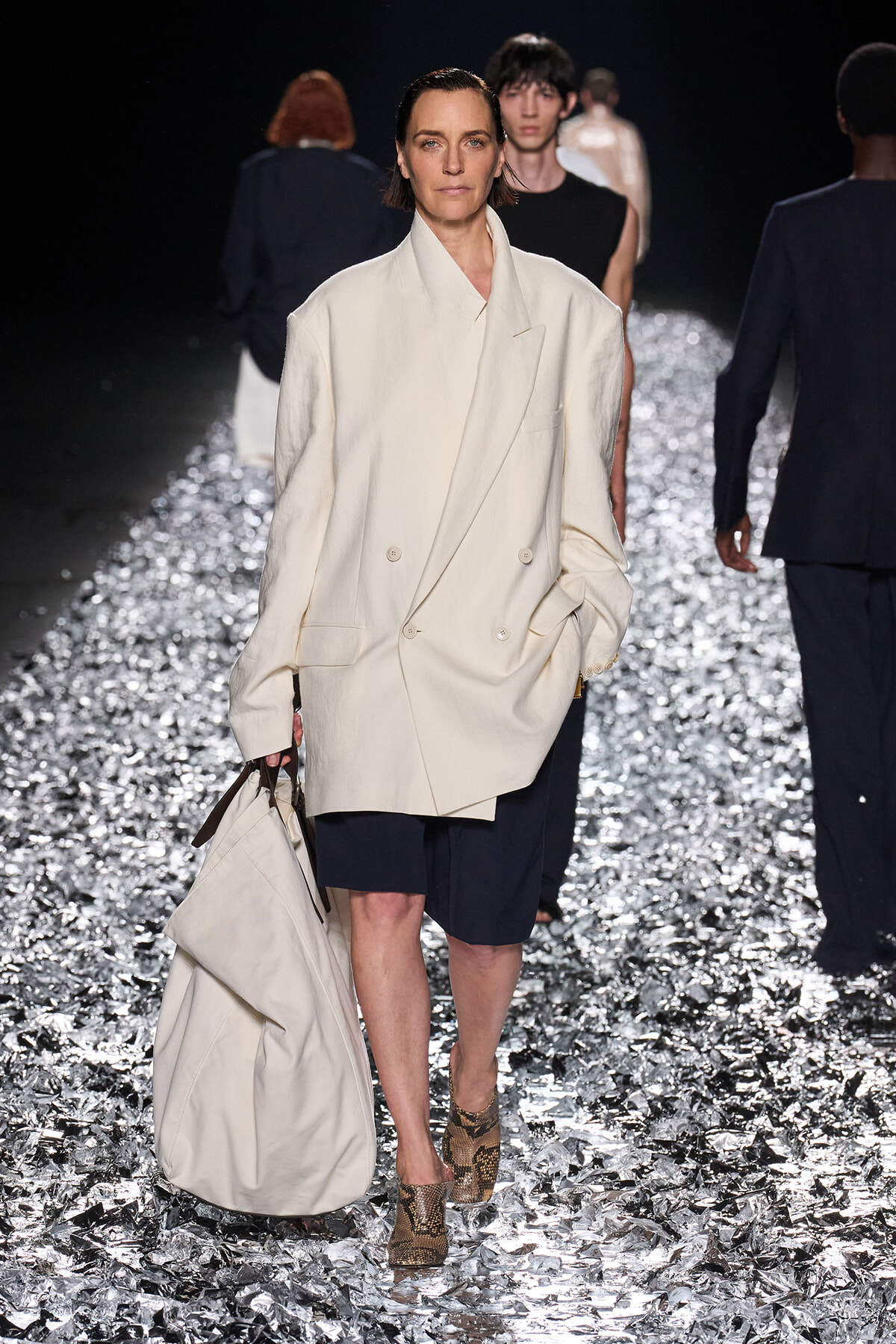 Runway model walking on a glittery catwalk in a cream oversized blazer, navy shorts, snakeskin boots, and a large light bag.