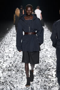 Model walks the runway in a navy windbreaker-style coat cinched with a wide brown belt, paired with black shorts and ankle boots on a reflective, crumpled metallic runway surface.