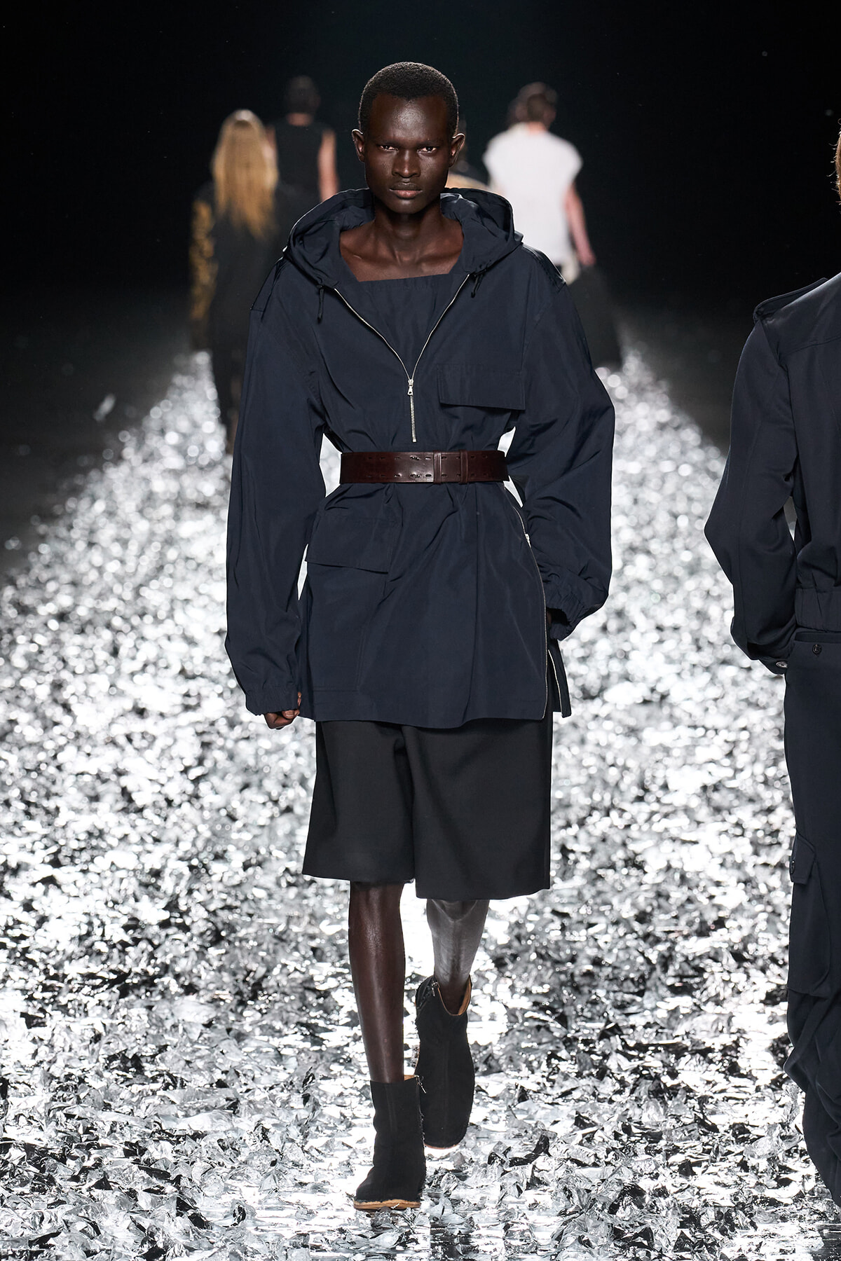Model walks the runway in a navy windbreaker-style coat cinched with a wide brown belt, paired with black shorts and ankle boots on a reflective, crumpled metallic runway surface.