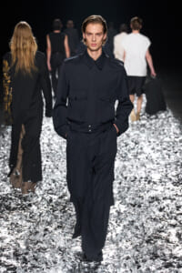 Male model wearing a navy utility jumpsuit with large front pockets, walking a glittering runway surface.