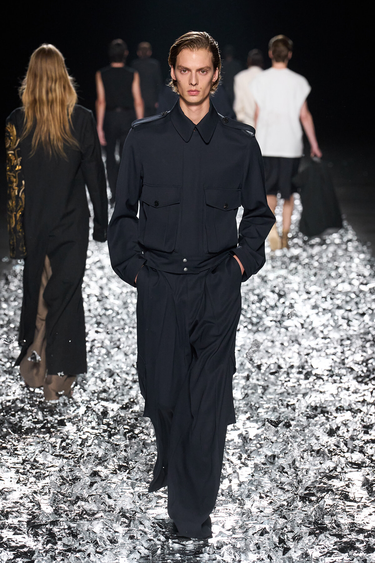 Male model wearing a navy utility jumpsuit with large front pockets, walking a glittering runway surface.