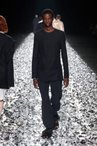 Male model in a black long-sleeve blazer and wide-leg pants walking a shiny silver runway in a fashion show.