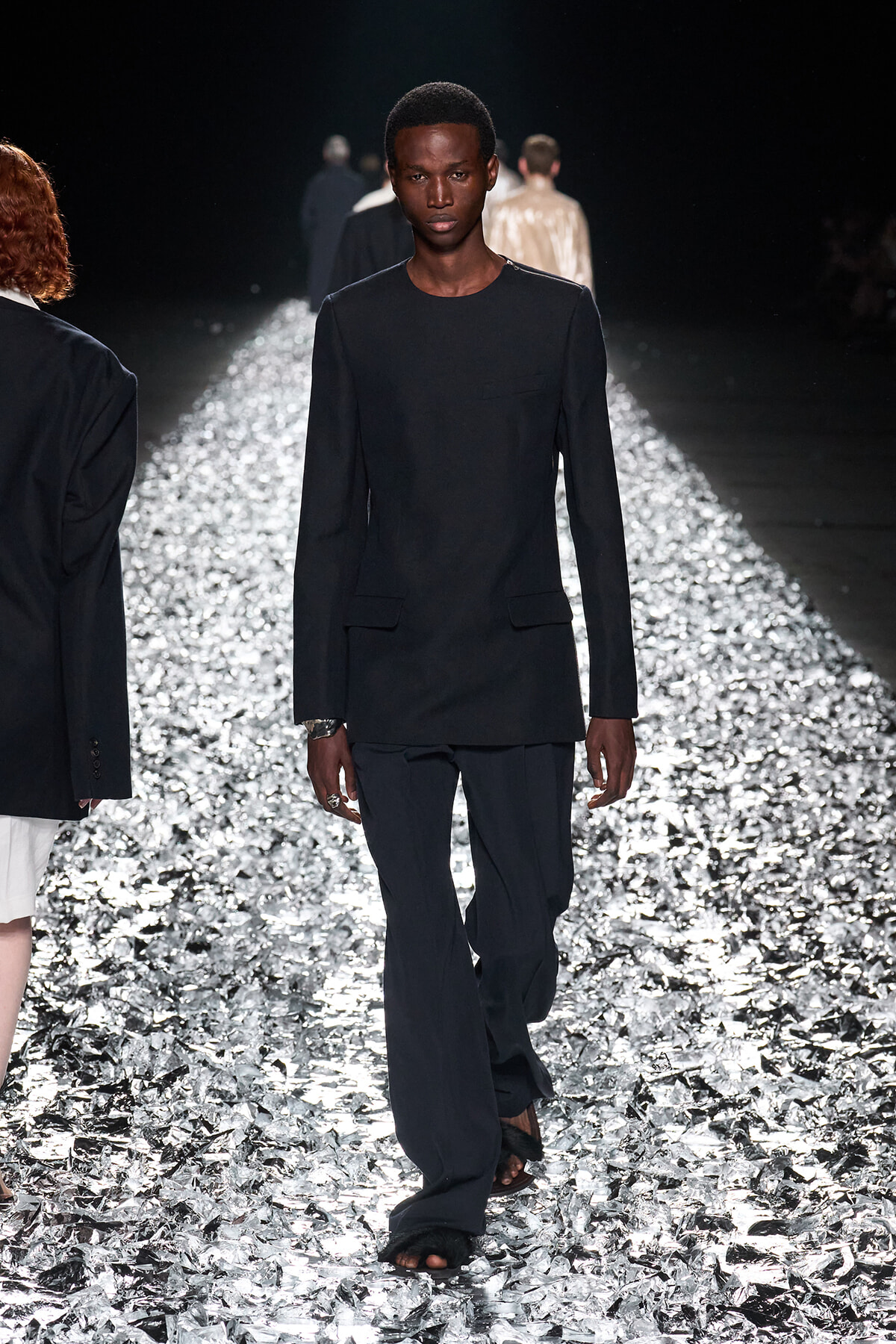 Male model in a black long-sleeve blazer and wide-leg pants walking a shiny silver runway in a fashion show.