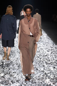 Model walks a sheer beige trench coat down a glittering runway, with other models blurred in the background.