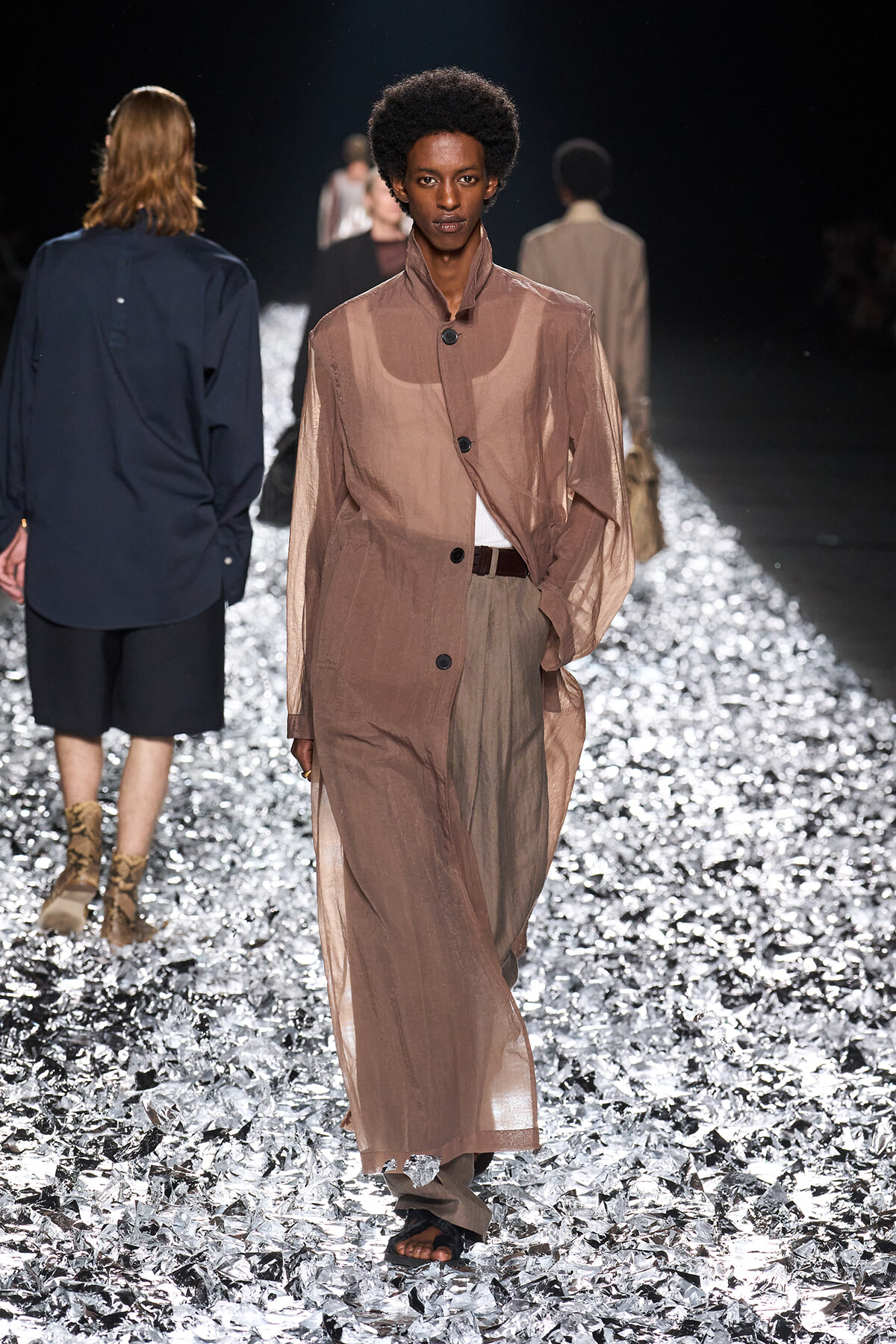 Model walks a sheer beige trench coat down a glittering runway, with other models blurred in the background.