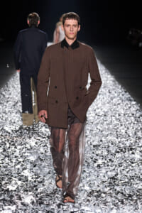 Male model walks a fashion runway in a brown pinstriped double-breasted blazer and sheer brown pants, barefoot sandals on a metallic foil floor.